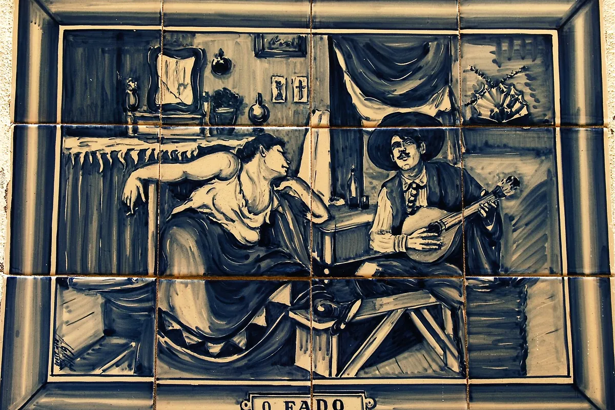 A blue and white tile panel showing a woman leaning on a table and a man in a hat playing a guitar, labelled O Fado.